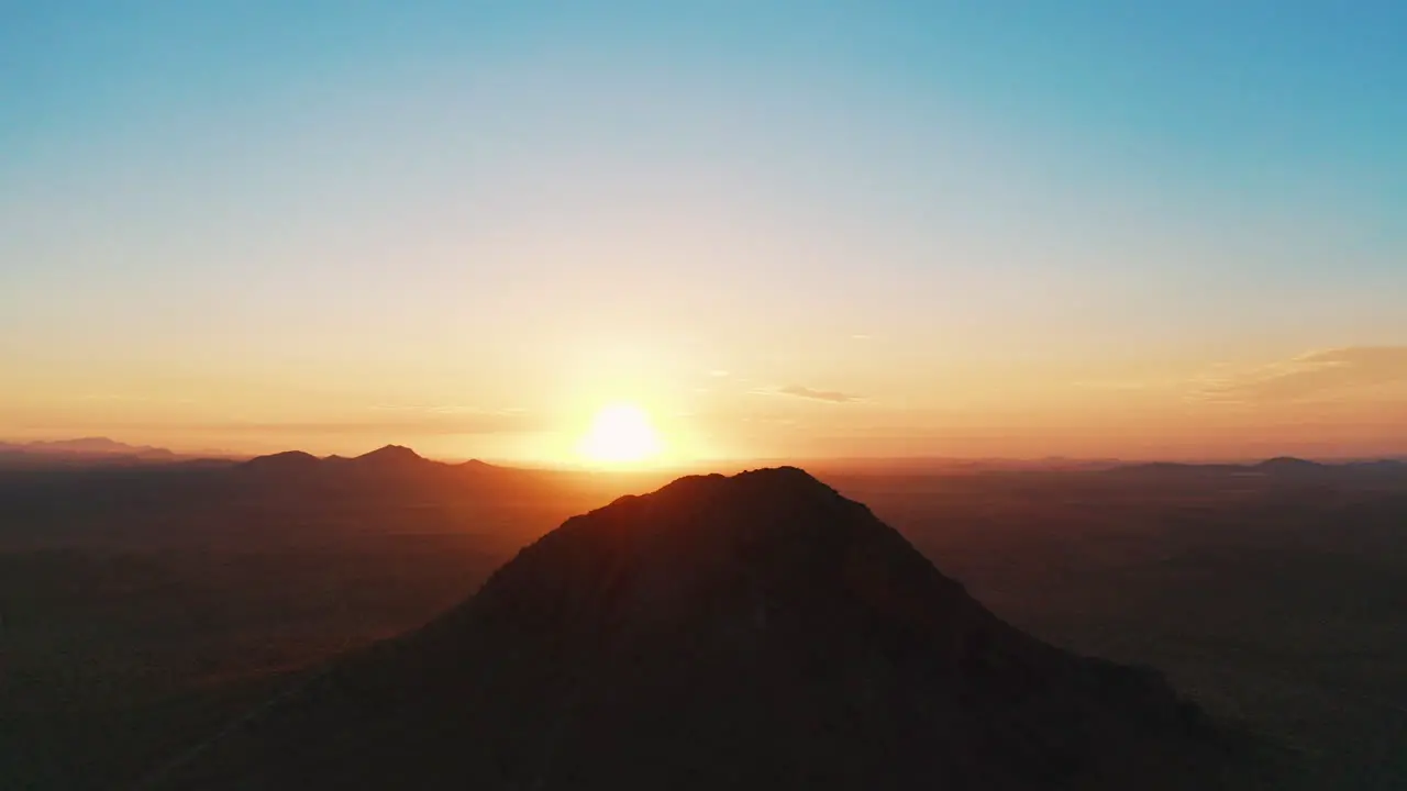 Slow aerial approach towards the rising sun glowing golden rays beyond a mountain peak in the Mojave Desert