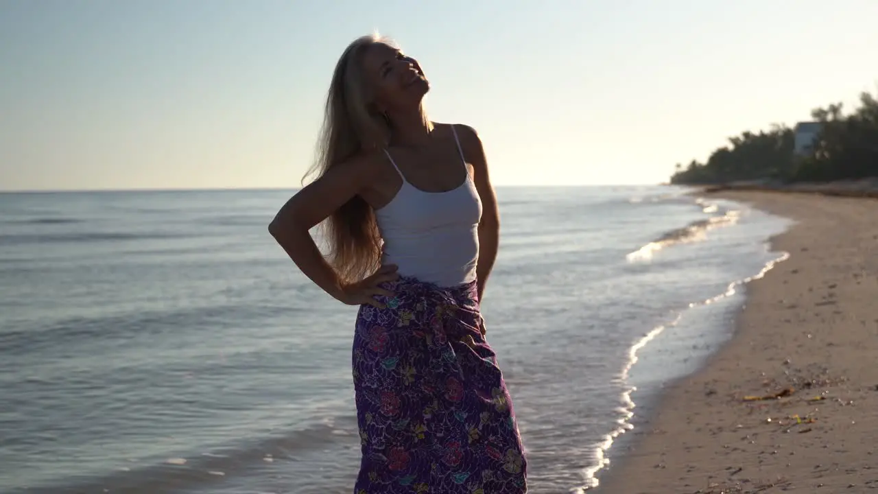 Pretty mature backlit woman on a beach looks away from the sea and then raises her arms and smiles looking to the sky