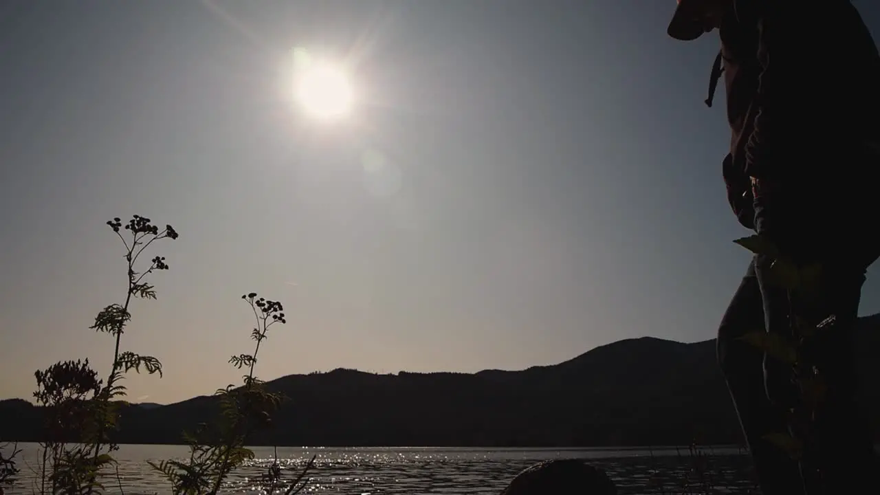 Silhouette of man looking at view with sun on shimmering lake with mountains in background