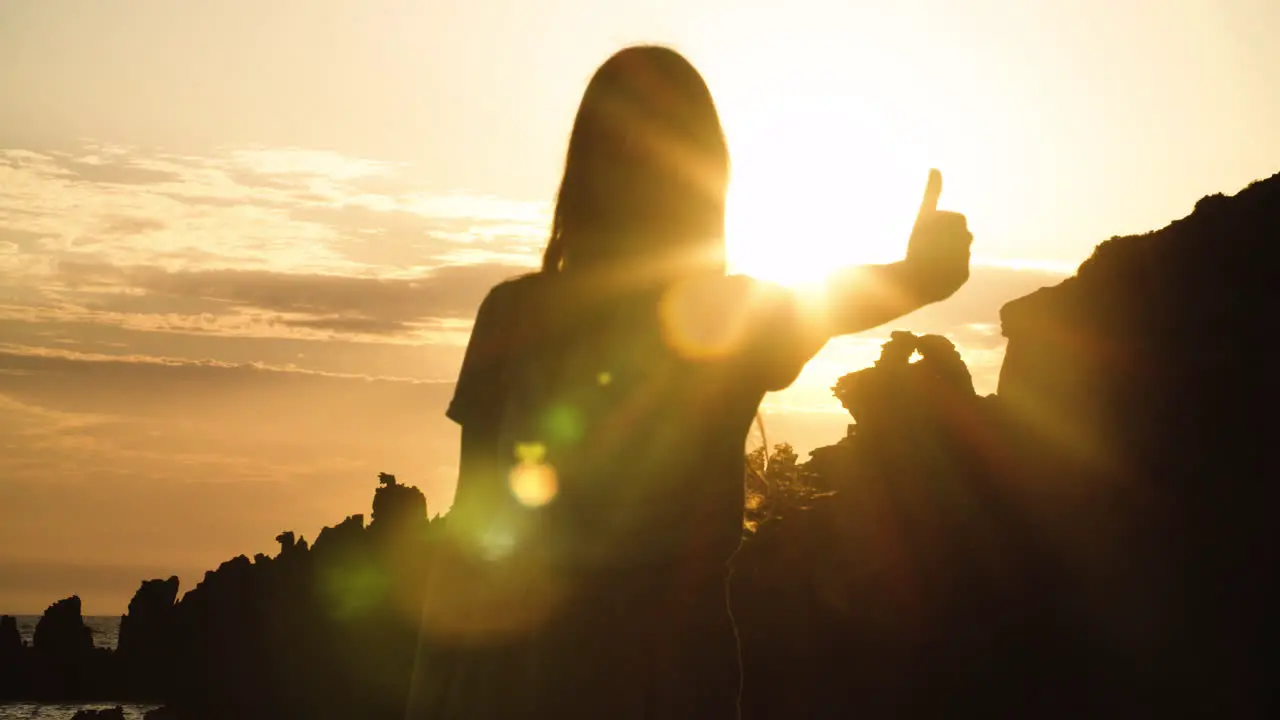 Young girl with long hair silhouetted by rocky landscape gives thumbs up with bright white sun and glare shining in background sky static