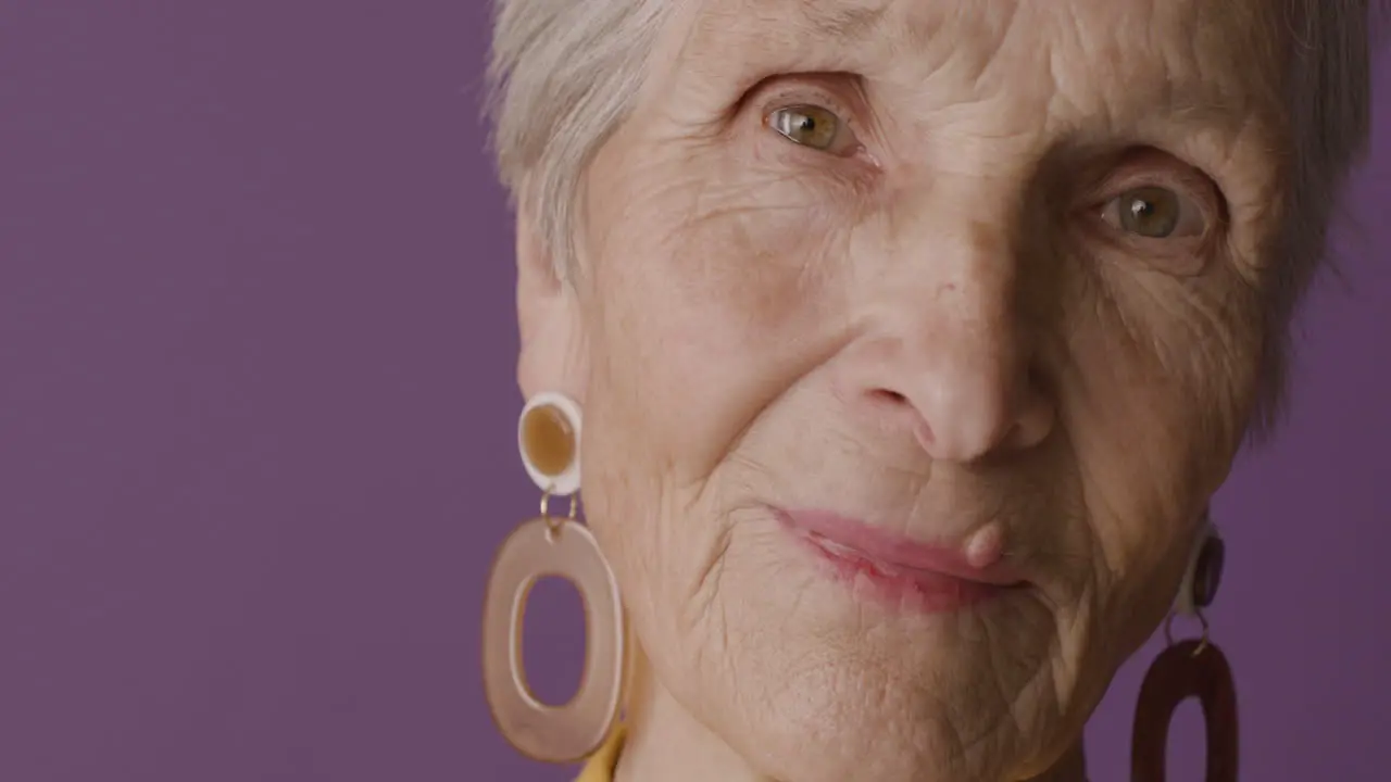 Close Up View Of Senior Woman Face With Short Hair And Green Eyes Wearing Earrings Posing At Camera On Purple Background