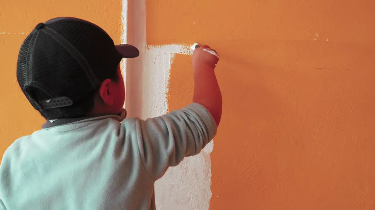 Child Wearing Cap Using Painting Brush To Paint Orange Wall Into White Color