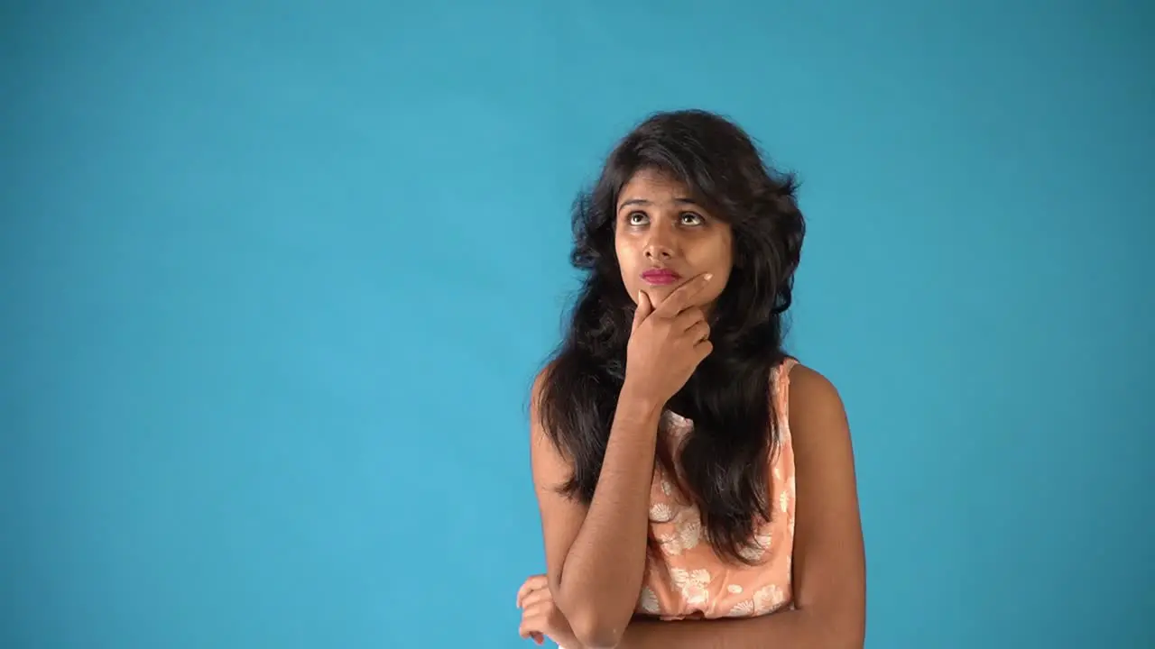 A young Indian girl in orange frock feeling worried and looking at the camera and then thinking in an isolated blue background