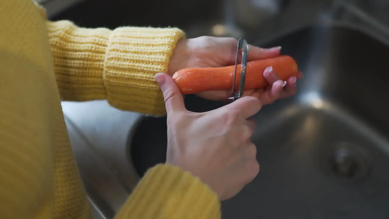 Closeup footage of a woman wearing a yellow sweater peeling a carrot with a peeler over the sink