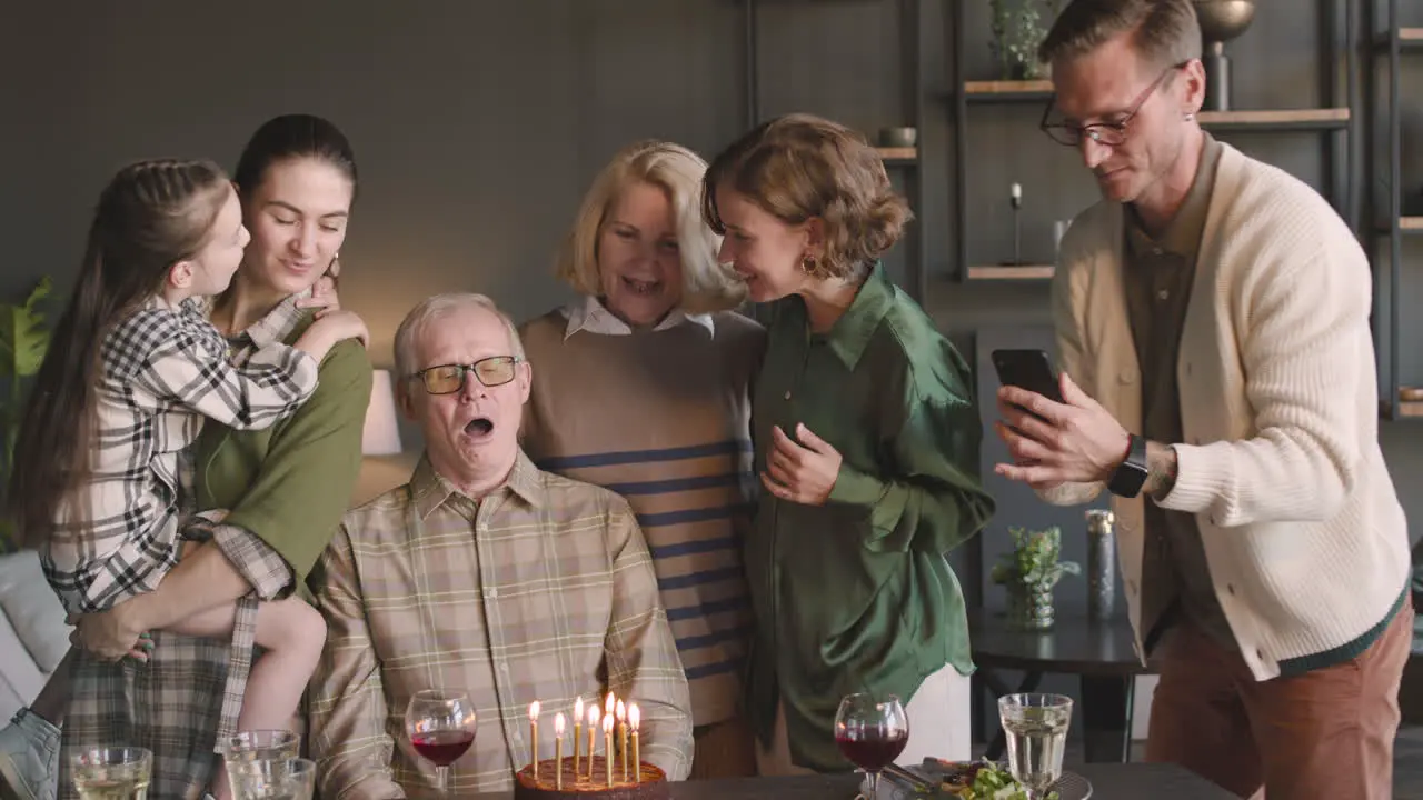 Senior Man Blowing Out Candles On Birthday Cake During A Celebration With His Family At Home While Relative Filming A Video With Mobile Phone