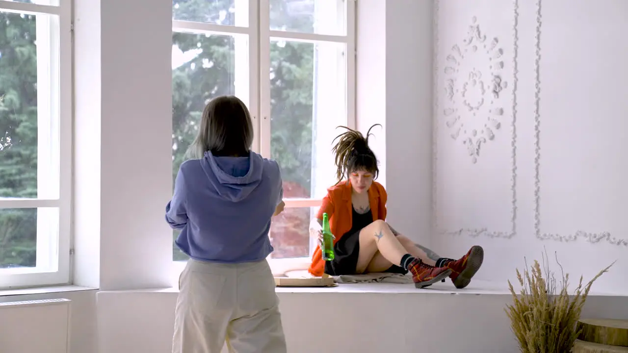 Brunette Girl With Dreadlocks Talking And Eating Pizza Next To The Window While Her Friend Approaches To Her With An Orange