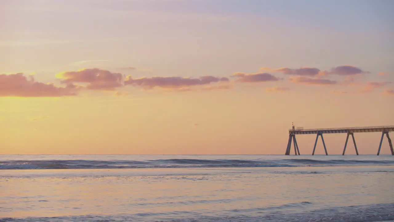 A dramatic orange beach sunset with light clouds and a jetty in the distance