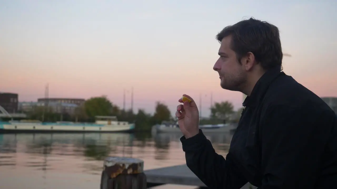 Wide shot of a young man snacking on an orange slice and smiling on a water pier during sunset