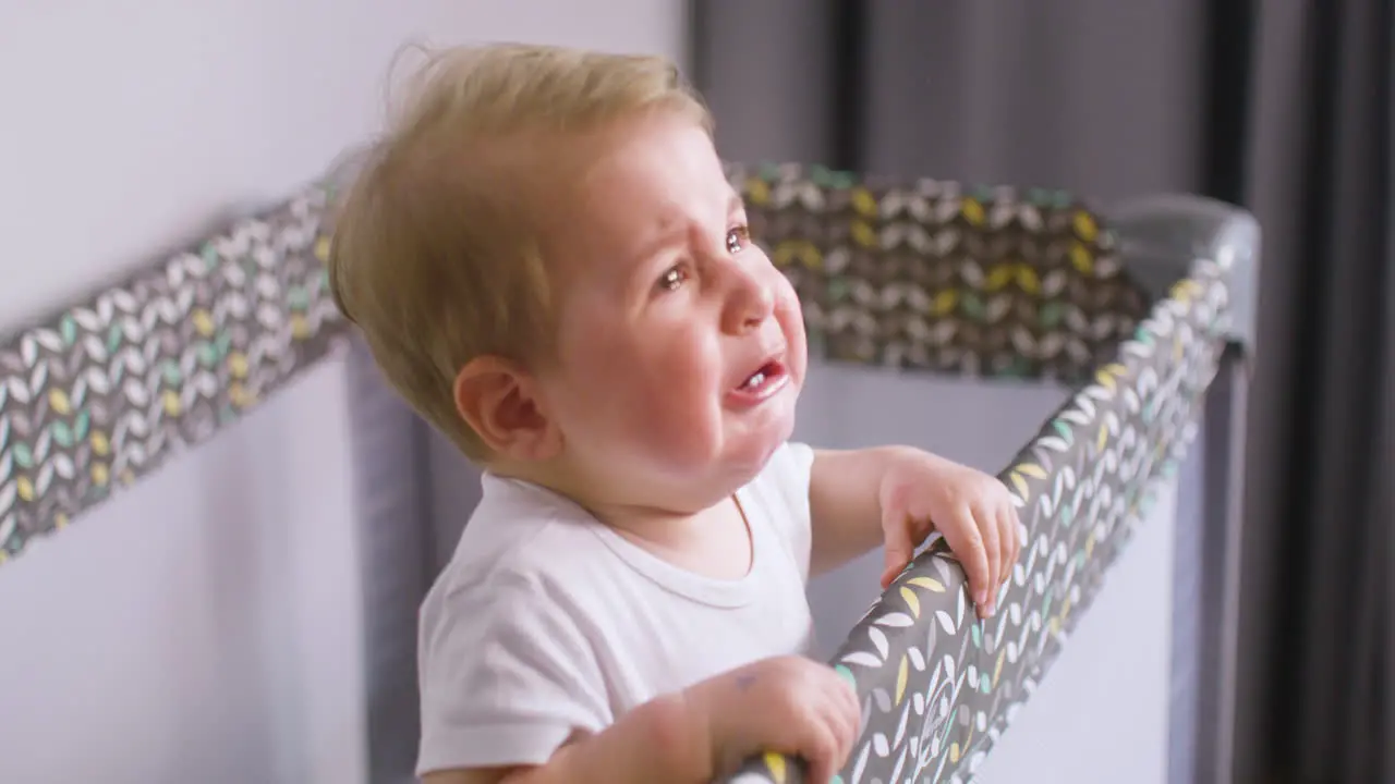 Side View Of Crying Baby In White Bodysuit Inside The Baby Playground While His Mother Giving Him A Fruit