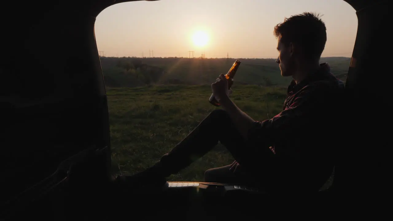 A Teenager Sits In The Trunk Of A Car And Drinks Beer From A Bottle