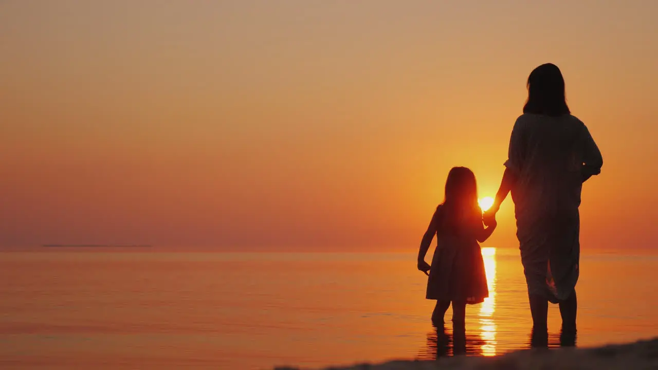 Silhouette Of A Pregnant Woman With A Baby Near By Stand Near The Sea At Sunset Waiting For The Seco