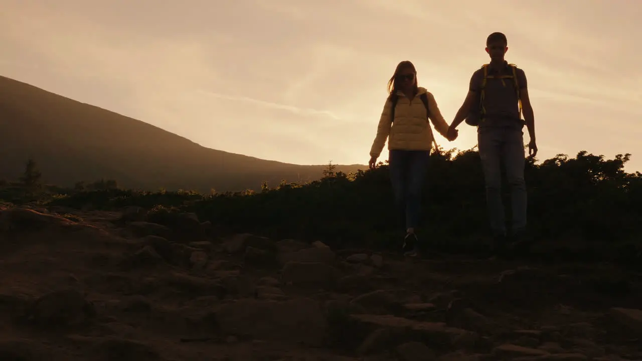 A Young Couple Of Tourists With Backpacks Walking Along A Mountain Trail In The Rays Of The Setting 