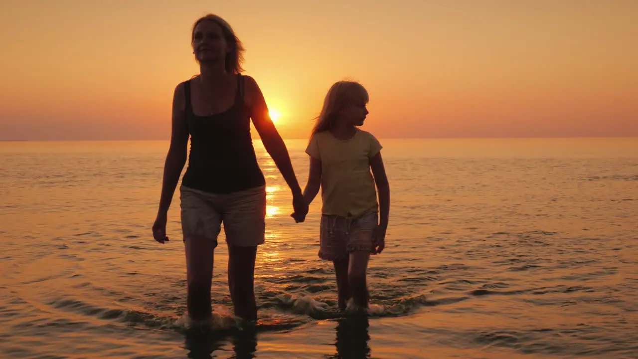 An Attractive Mother Walks With Her Daughter On The Water Against The Backdrop Of The Setting Sun A 