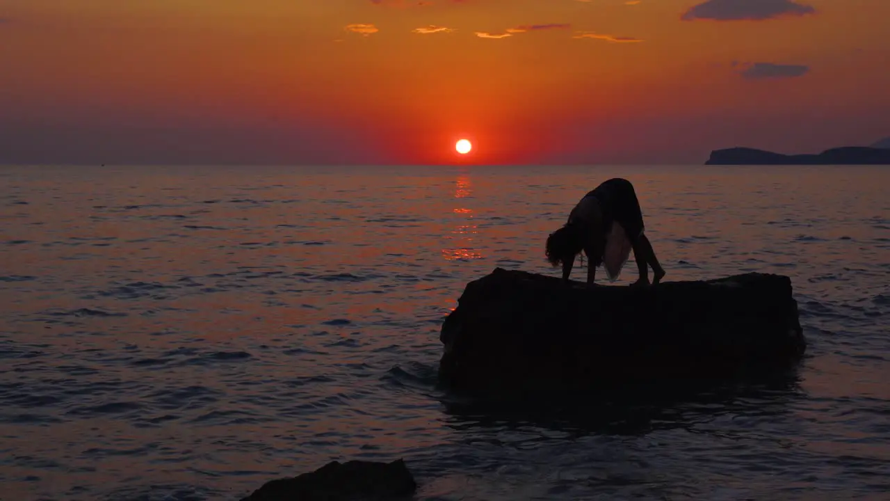 Young woman doing Yoga exercises on big cliff surrounded by sea water reflecting red sky of sunset