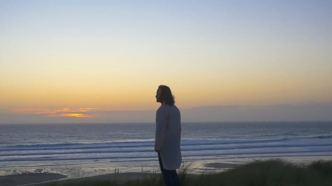 Man in kaftan stands on beach watching ocean sunset wide