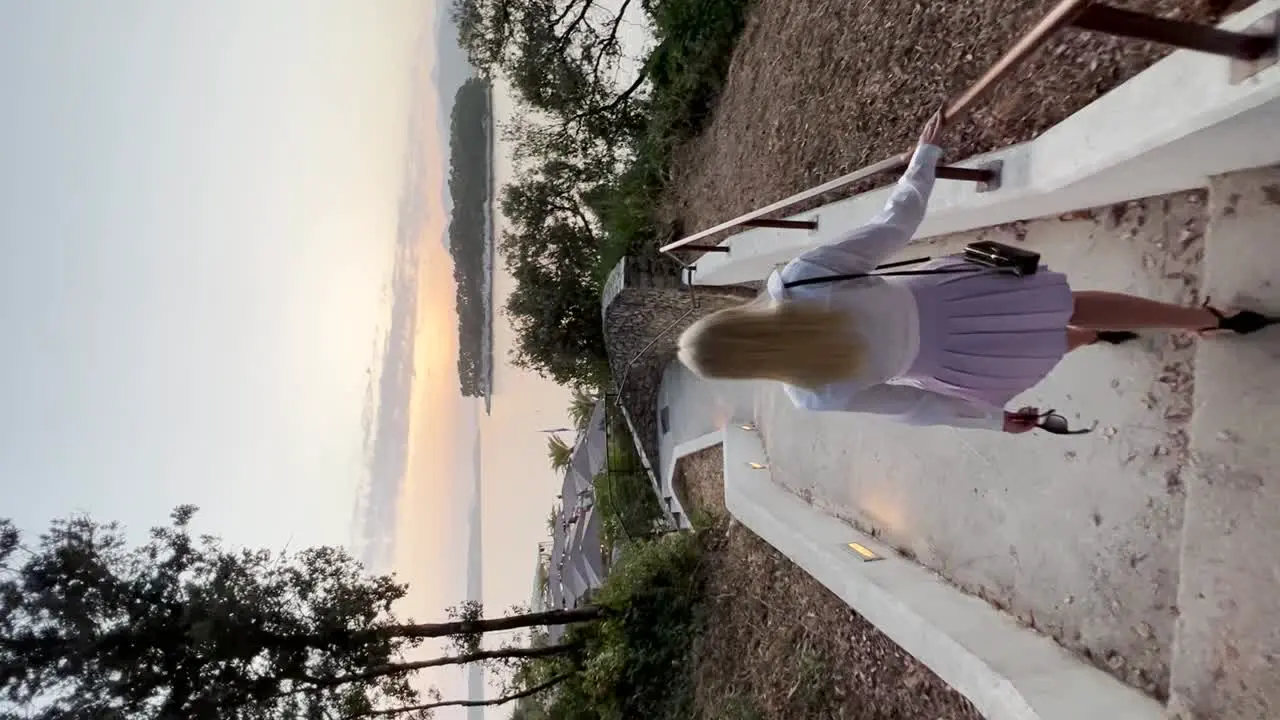 A young woman dressed in a white shirt and purple skirt walks down the white staircase to the beach club with a sunset over the beautiful Adriatic ocean in Dubrovnik