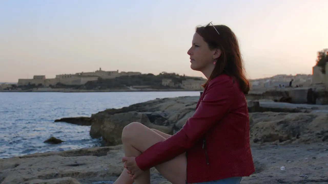 Young Girl With Red Hair Seats On Rocky Shore Viewing Seascape During Sunset