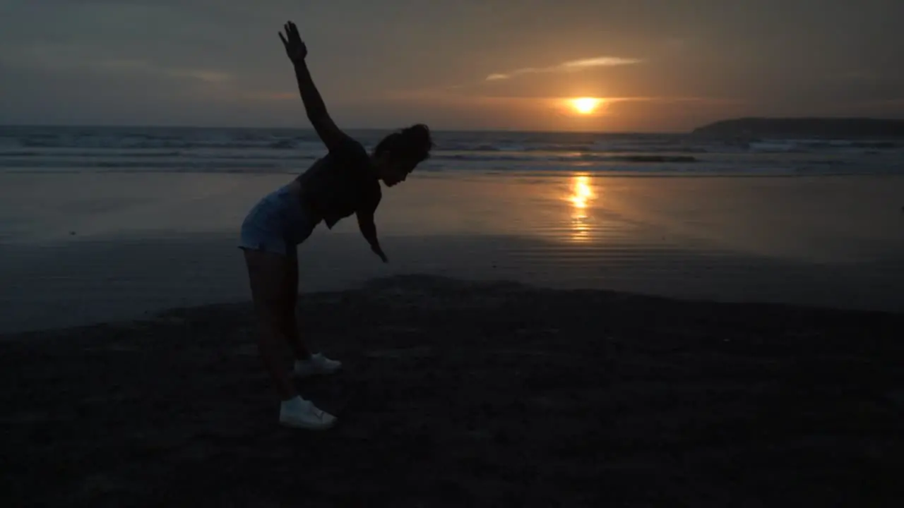 Slow motion landscape view of a young woman silhouette doing stretching exercises on a sandy beach by the ocean at sunrise