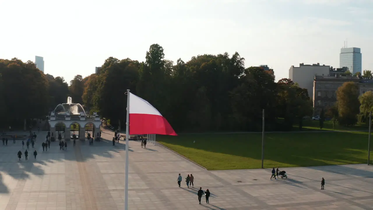 Orbit shot around national flag of Poland waving on pole on Pilsudski Square People walking across tiled square of monument in rest of Saxon Palace Warsaw Poland