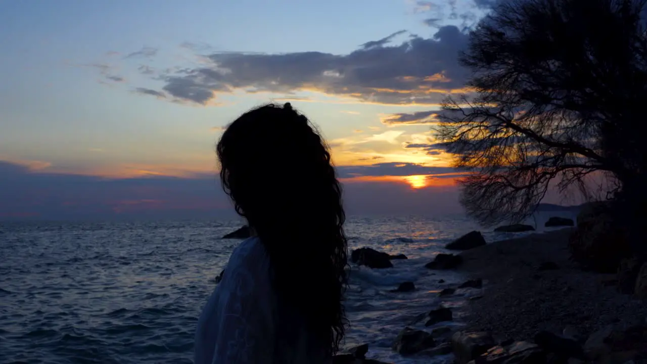 Girl meditates on rocky beach watching beautiful sunset with cloudy sky reflecting on vibrant sea water