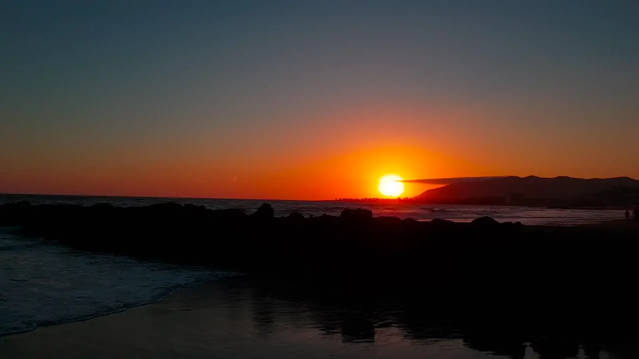 Slow and low shot rising above rocks and revealing sunset with silhouettes of people by the shore at San Buenaventura State Beach in Ventura California United States