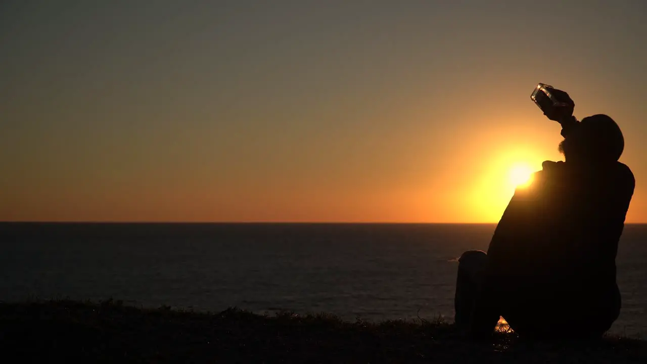 Silhouette of a desperate man drinking from the bottle while sitting on the beach at sunset with copyspace