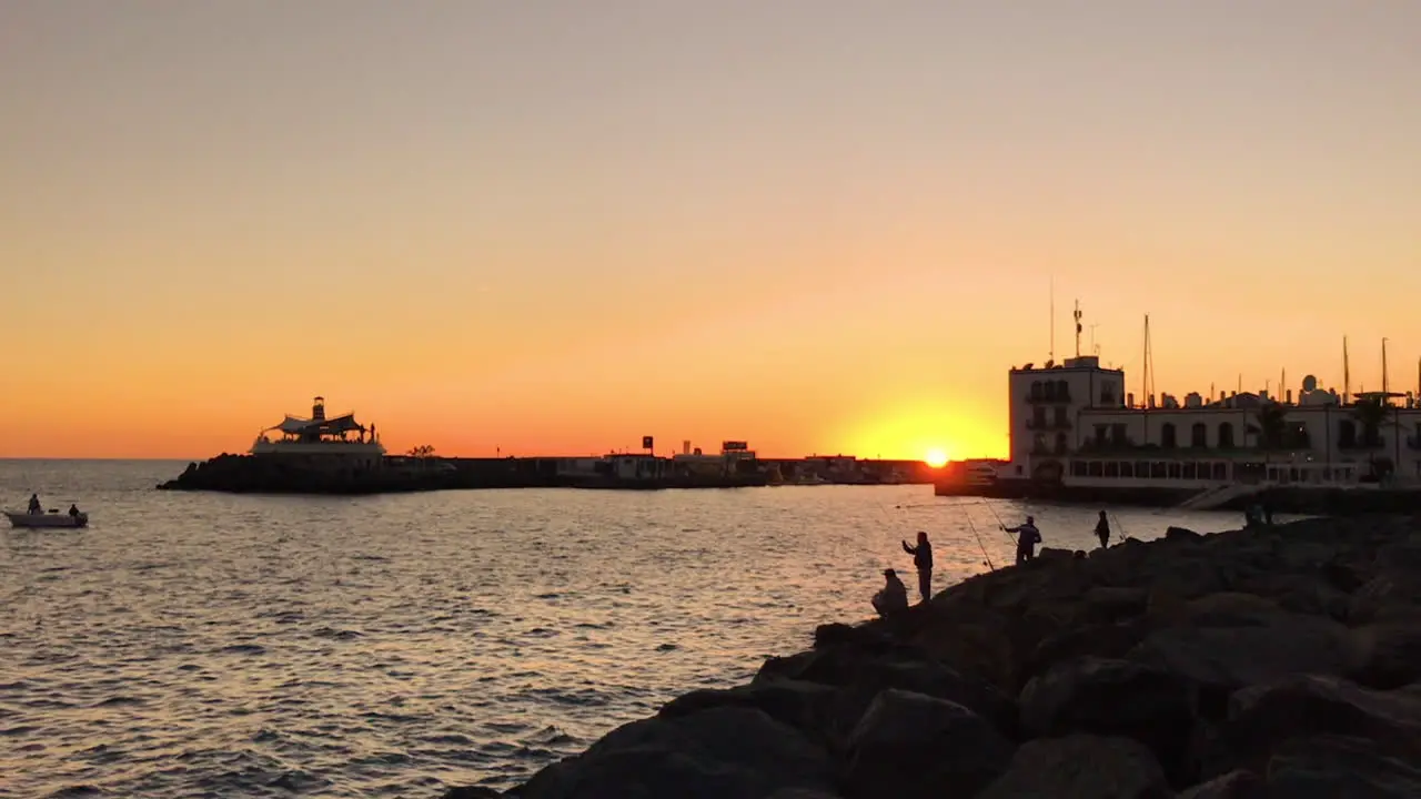 Silhouettes of fishermen on pier catching fish during orange sunset casting fishing line on open sea in background of bay and part of city in slow motion capture at 120fps