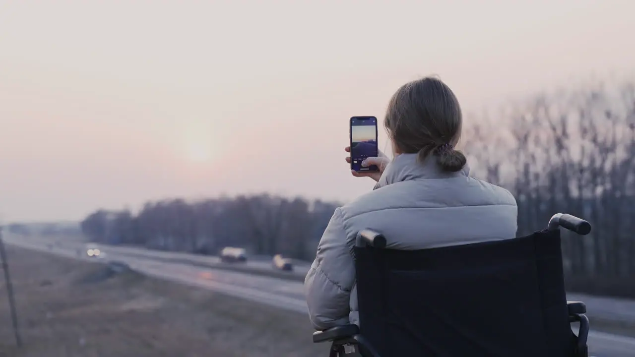 Rear View Of Disabled Woman In Wheelchair Recording The Landscape At Sunset