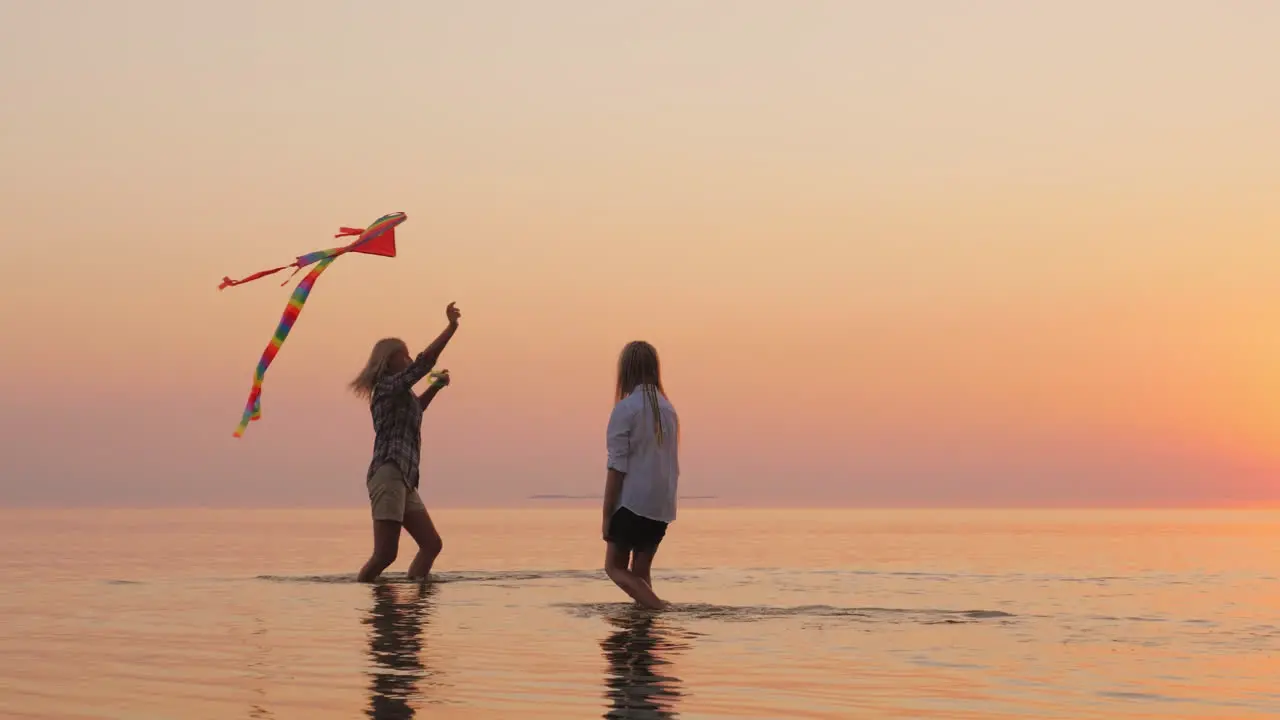 Mom And Daughter Are Playing With A Kite At Sunset On The Water Of A Beautiful Lake