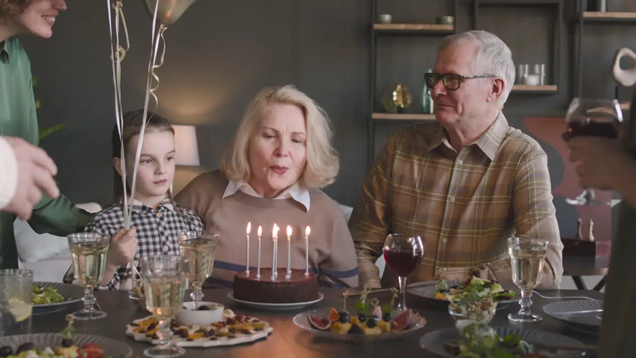 Senior Woman Blowing Out Candles On Birthday Cake During A Celebration With Her Family At Home