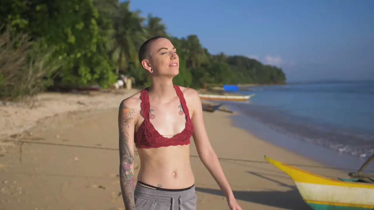 Thin woman with shaved head standing on a beach at the edge of the water with her face to the sun