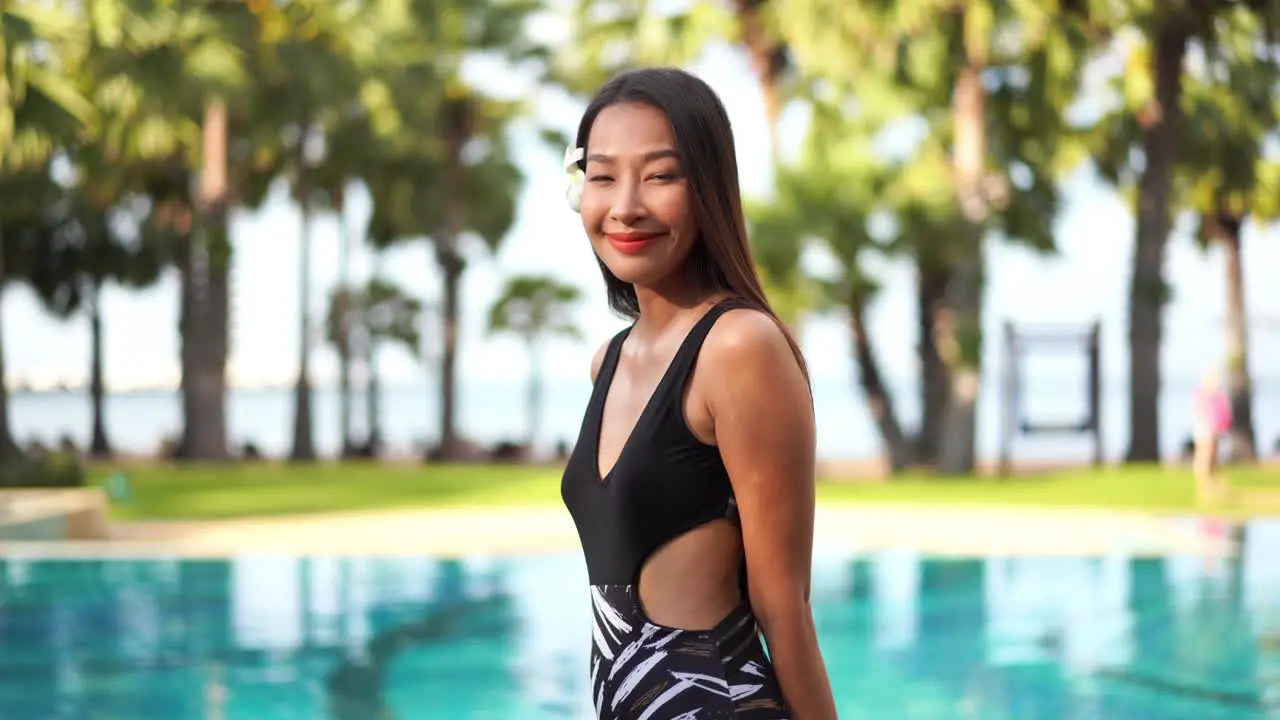 Beautiful shy Asian girl with black bathing suit smiling to the camera blurred swimming pool background