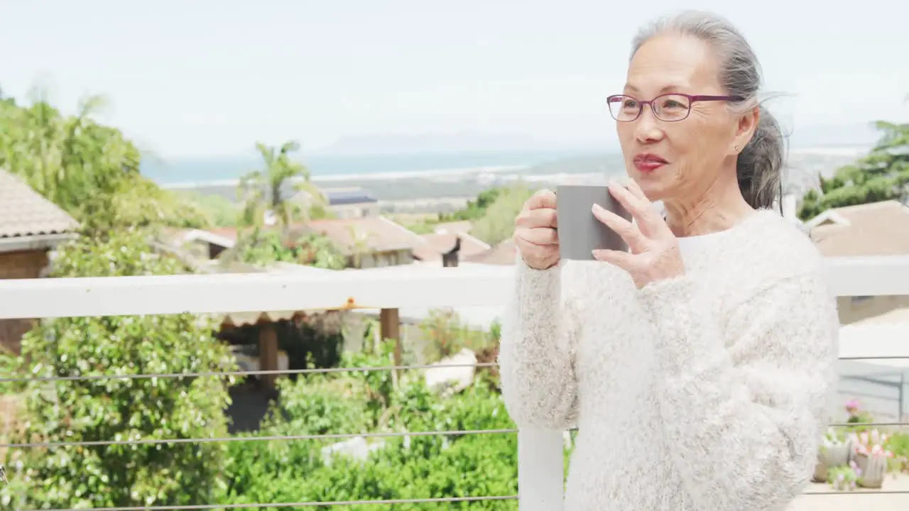 Happy asian senior woman relaxing on balcony with mug of coffee