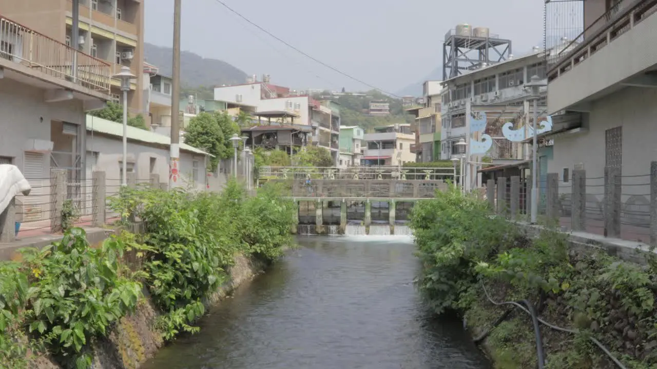 A river passing through a canal with 3-story buildings on both sides and a small dam bridge in the background in a small village in southern Taiwan wide shot