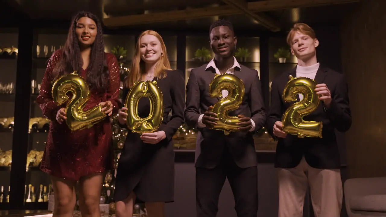 Group Of Friends Wearing Elegant Clothes Holding Balloons With The Numbers 2022 While Dancing In New Year's Party