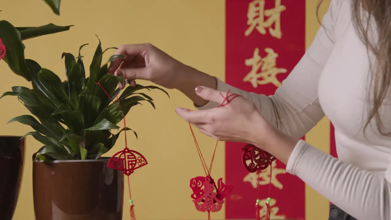 Woman Hanging Decorations Celebrating Chinese New Year On Plants At Home With Banner In Background 1