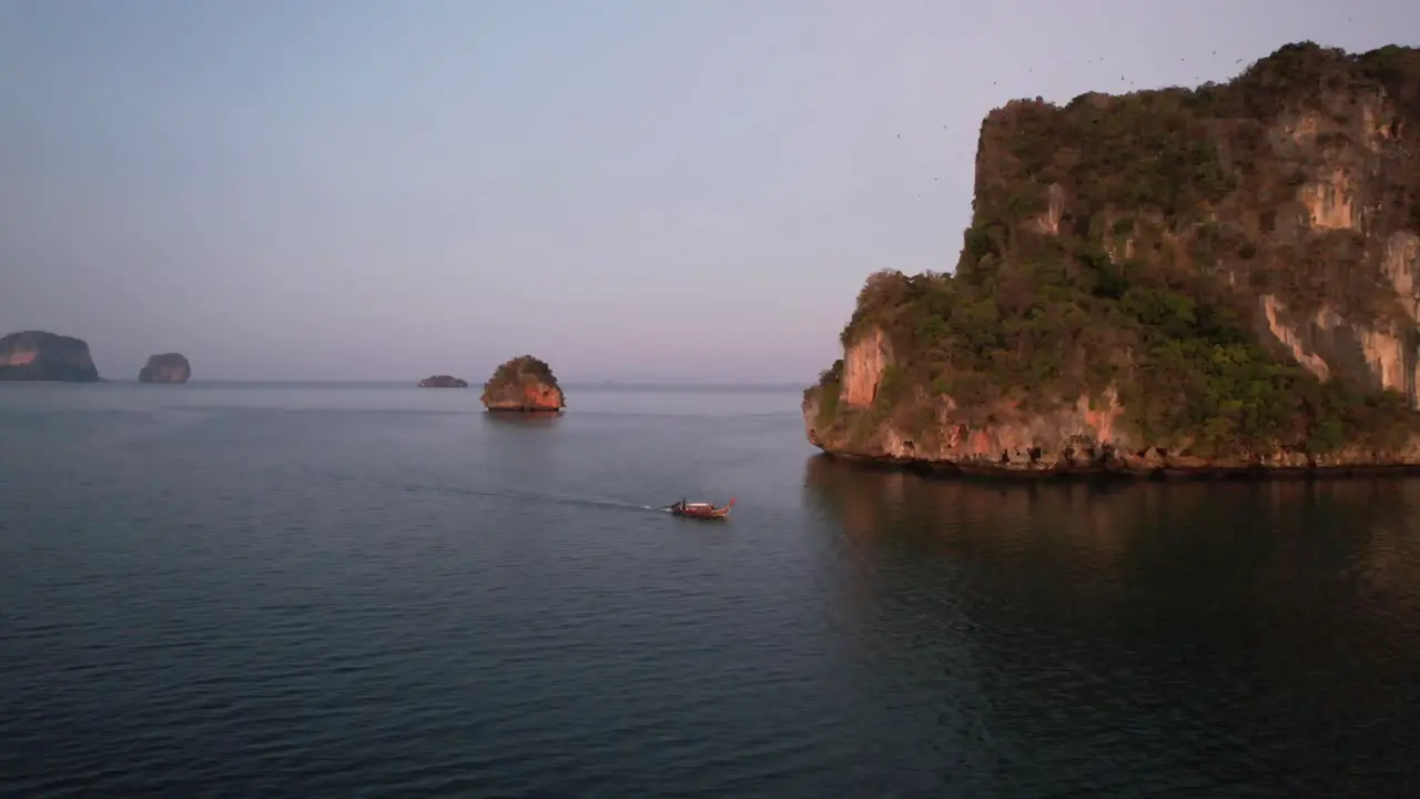 wide aerial shot of a single thai longtail boat motoring near large limestome mountains of Railay Beach in Krabi Thailand during sunrise in the Andaman Sea