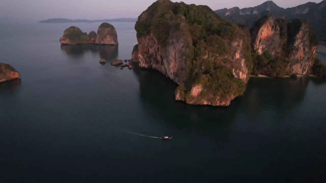 Wide high altitude aerial drone of unique limestone mountain formations near Railay Beach in Krabi Thailand during sunrise as a thai longtail boat motors in the Andaman Sea