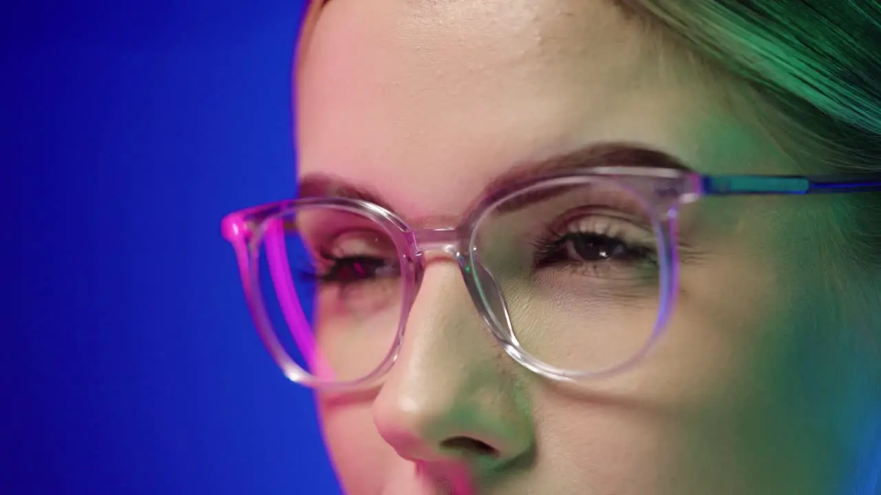 Slow motion close up shot of a pretty attractive woman wearing glasses while she adjusts the glasses with her finger on her face with purple light in her face against blue background