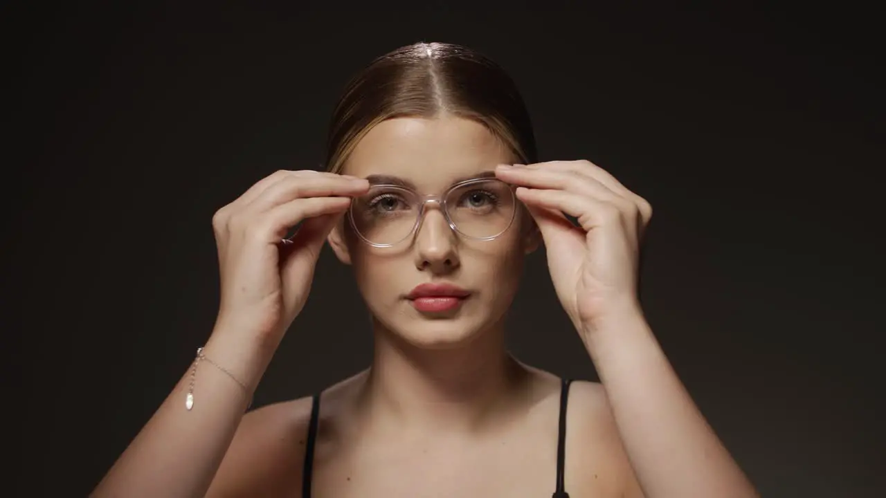 Medium shot of a young pretty woman who needs a visual aid because her eyesight is not good enough and puts on glasses and looks to the camera in front of black background in slow motion