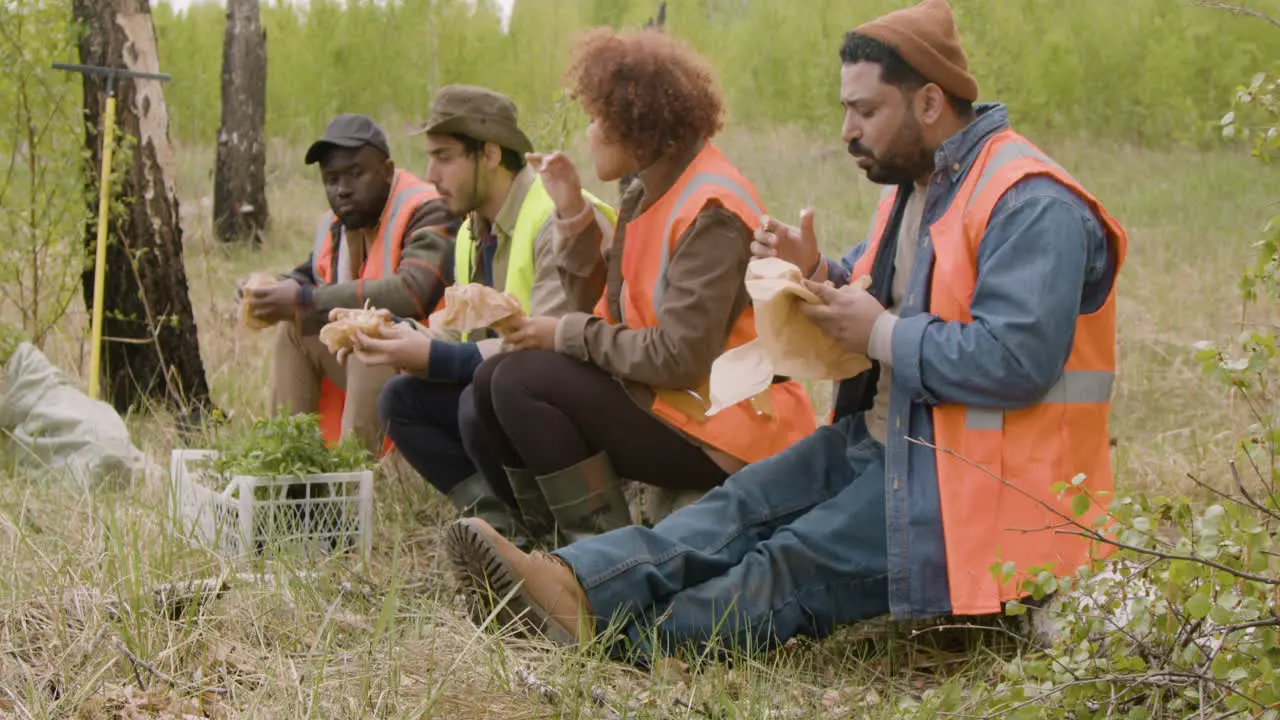 Side view of a group of multiethnic ecologist activists eating and talking in a break sitting on a trunk in the forest