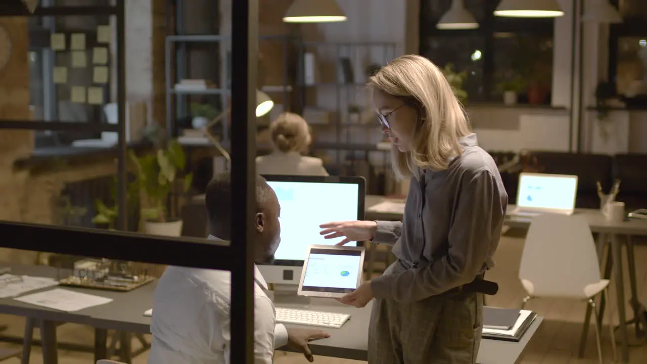 Rear View Of American Man Sitting At Desk While Talking With Female Coworker Who Is Showing Him Graphics On The Tablet Device In The Office