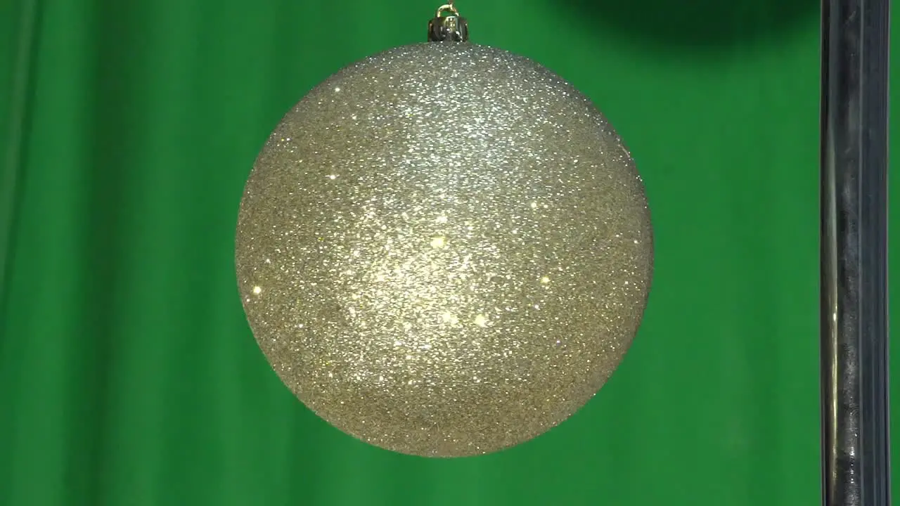 This video shows a gold glitter Christmas ornament ball hanging by a string and slowly spinning around in front of a green screen