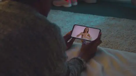 Woman Lying On Bed At Home Looking At Beauty Product Demonstration Video On Mobile Phone