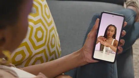 Close Up Of Woman Sitting On Sofa At Home Looking At Beauty Product Demonstration Video On Mobile Phone