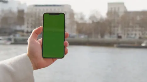 Close Up Of Man Holding Green Screen Mobile Phone Looking Out To River Thames And Embankment In London UK