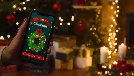 Hand At Home Holding Mobile Phone With Merry Christmas Message And Tree And Presents In Background