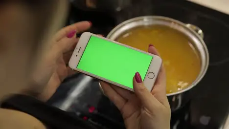 Woman is holding a smartphone with a green screen and cooking soup