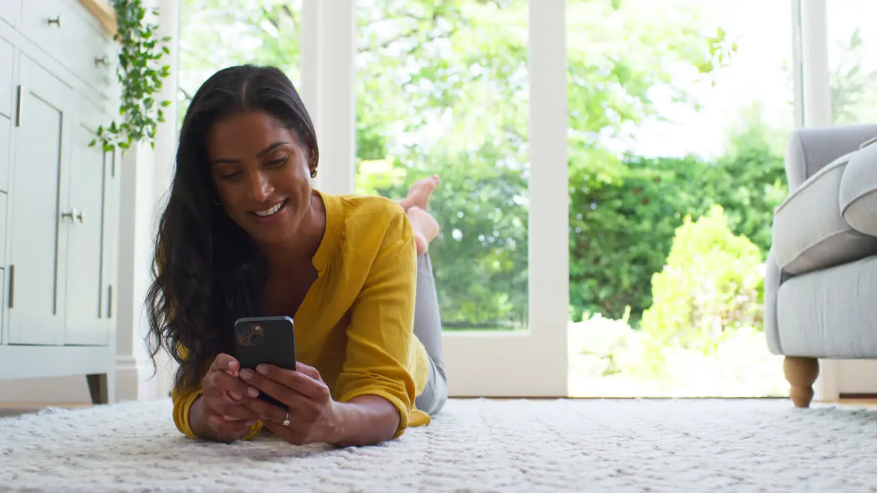 Woman At Home Lying On Floor In Lounge Streaming Movie To Mobile Phone