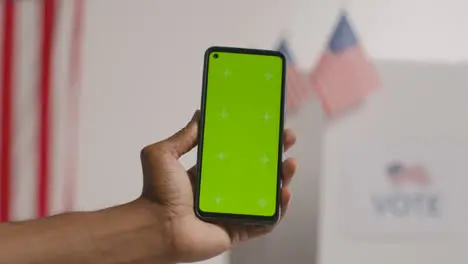 Close Up Of Hand Holding Green Screen Mobile Phone In Front Of Ballot Box In American Election